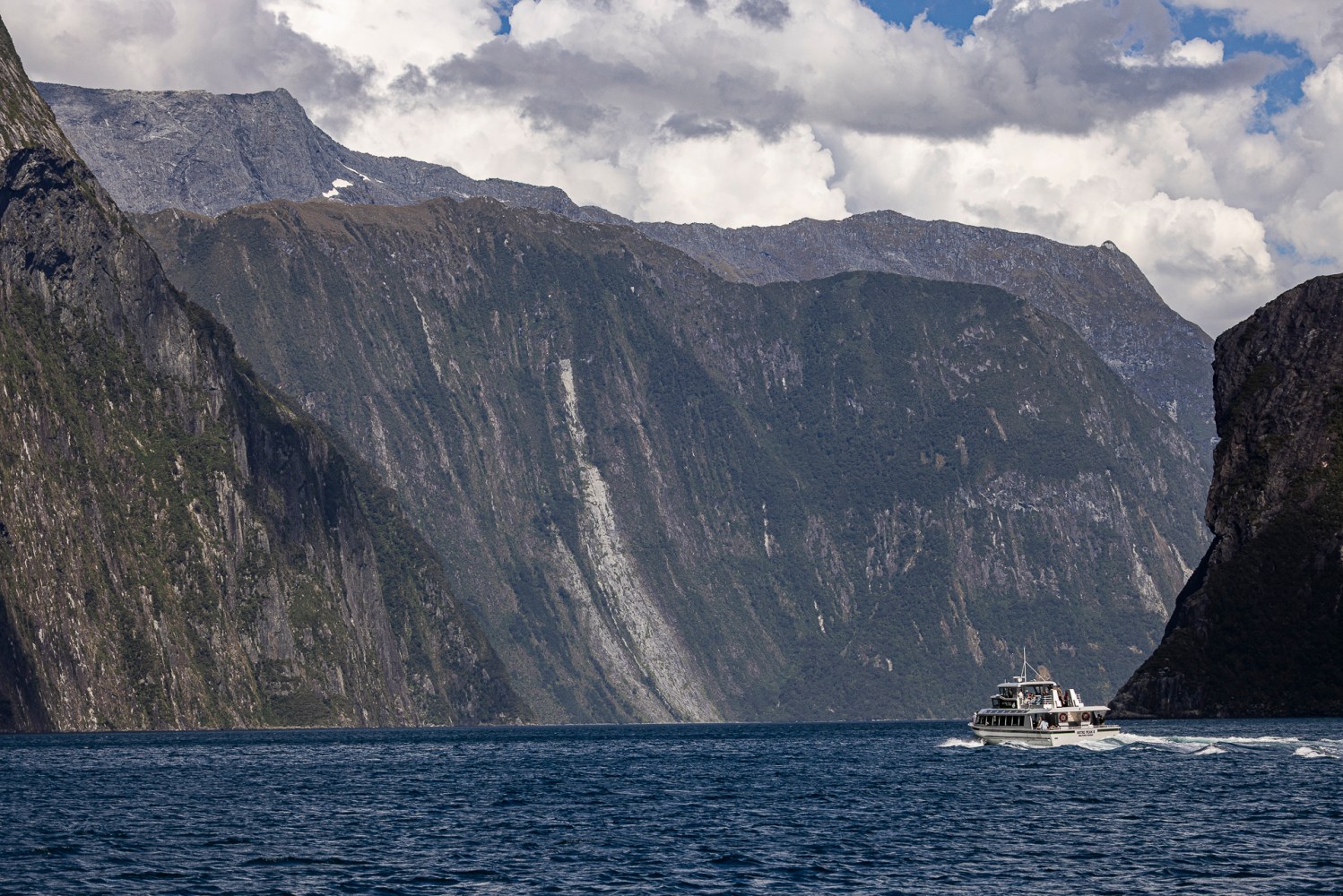 Boat on a fjord with steep cliffs and cloudy sky in the background.